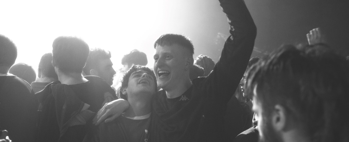 Medium black-and-white shot of two young men at an underground rave, laughing with their arms around their shoulders.