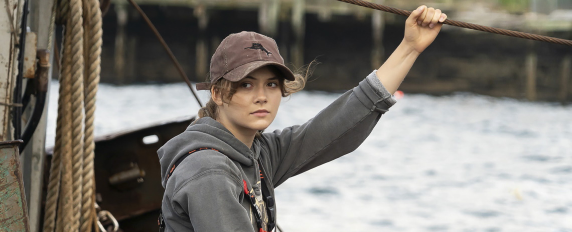 A medium shot of a teenage girl in a grey hoodie and ball cap, standing on a fishing boat.