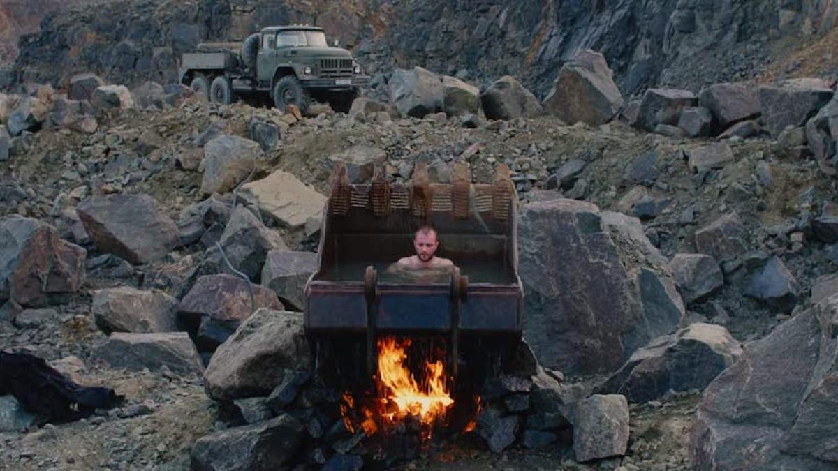 An unclothed man sits in a freestanding backhoe bucket filled with water. A fire is burning underneath the bucket. The surrounding landscape is gray, rocky, and barren. A water trucks is parked in the background.