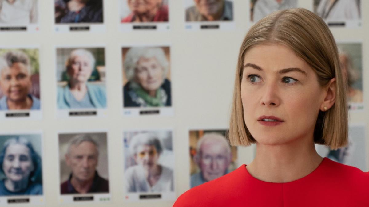 A woman with a blonde bob haircut and red shirt, standing in front of a wall of headshot photographs of elderly people.