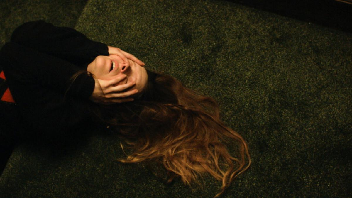 A woman in a black and red sweater lays on a green carpeted floor, her hands partly covering her face and her hair spread out on the ground.