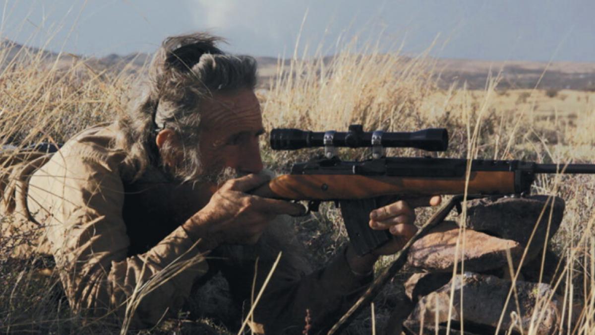 An older, bearded man laying down in a field of dry grass, pointing a rifle with a scope at something offscreen.