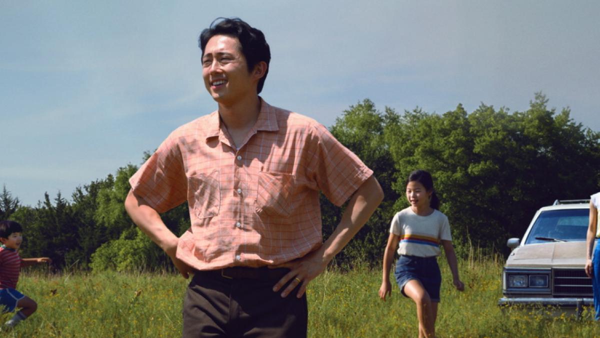 A man in a red-and-white checked shirt standing with his hands on his hips, surveying a grass field. His family -- wife, daughter, and young son -- are in the background with the family car.