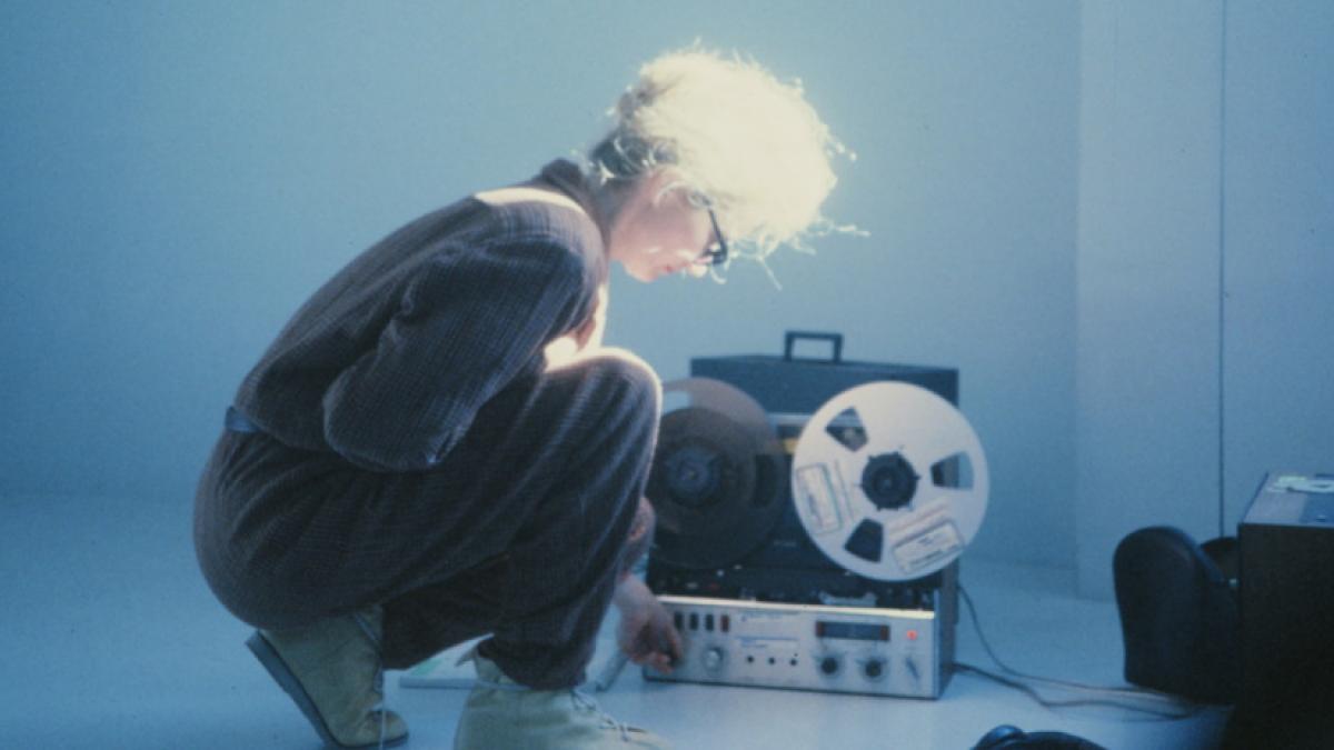 A woman crouches in profile, inspecting a real-to-real audio device.