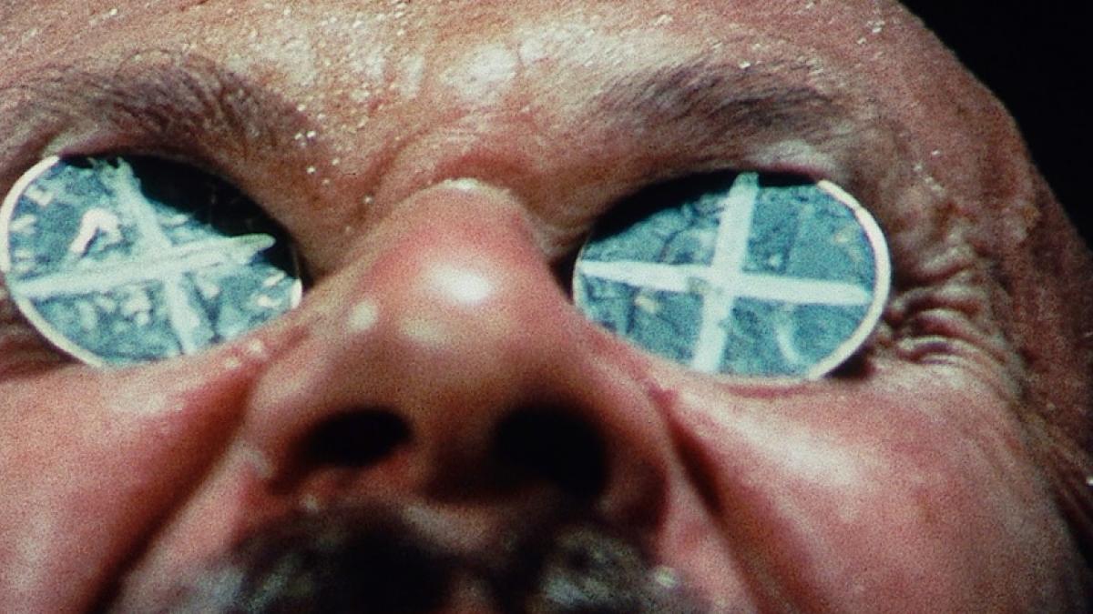 A close-up of a man's sweaty, upturned face. Two pennies painted with X's have been placed on his eyes.