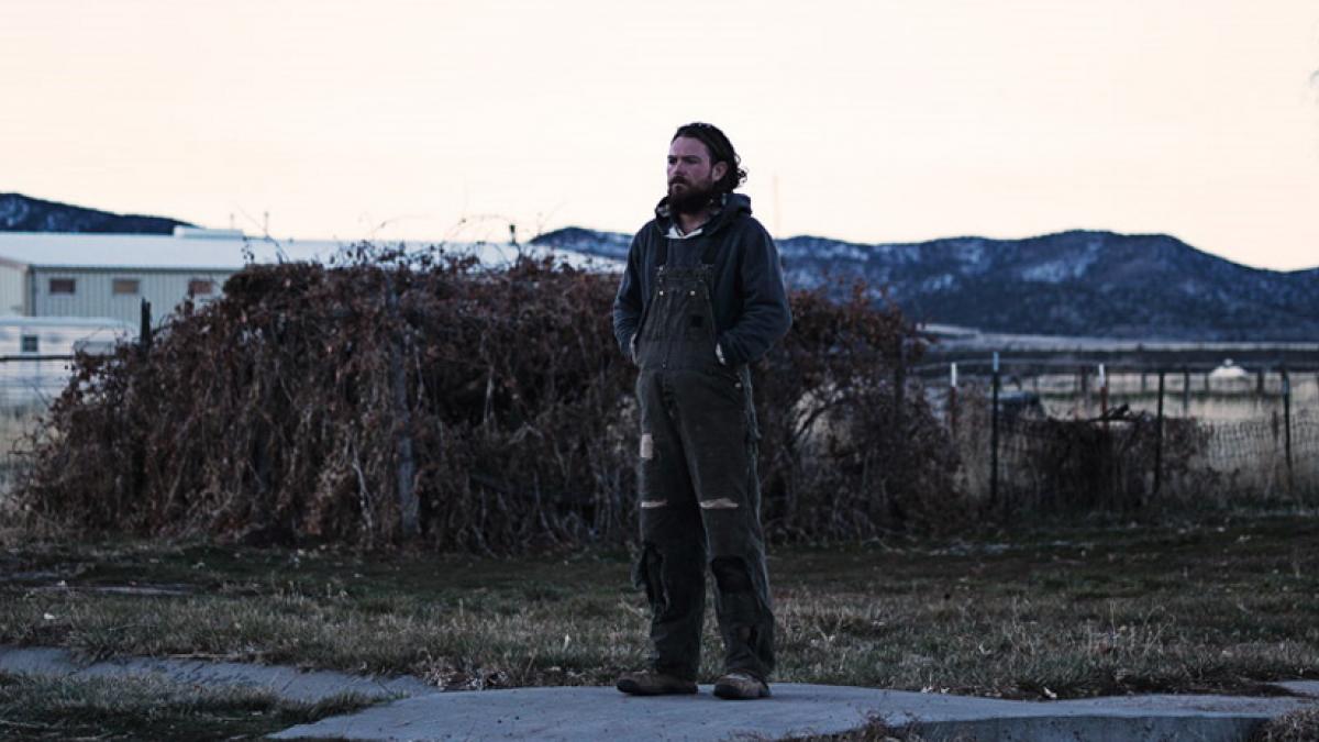 A bearded man in a hoodie and coveralls standing in a vacant lot in front on an overgrown fence, with mountains in the distance.