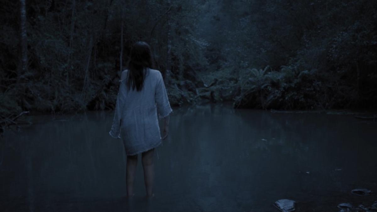 A woman in a white shift stands in a forest pool up to her knees, her back to the camera.