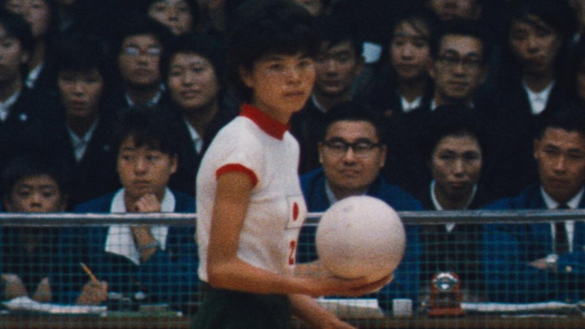 A vintage photo of a volleyball player holding a ball, about to serve. In the background is an audience of spectators.