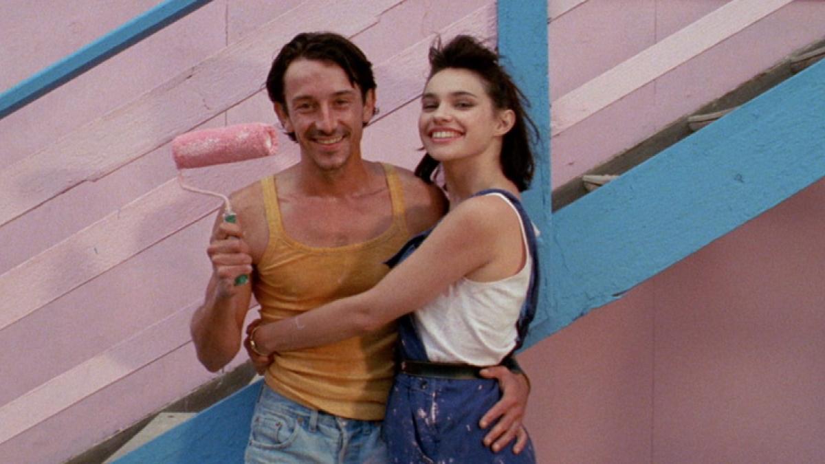 A shirtless man holding a paint roller covered in pink paint poses with a young woman in overalls in front of a wooden building painted pink and blue.