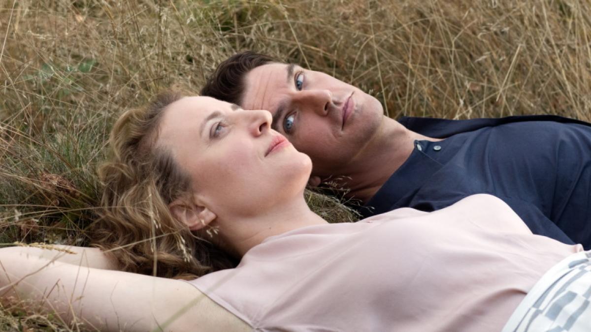 Medium shot of a middle-aged woman laying in the grass with a slightly younger man; she is looking up into the sky, he is looking at her.