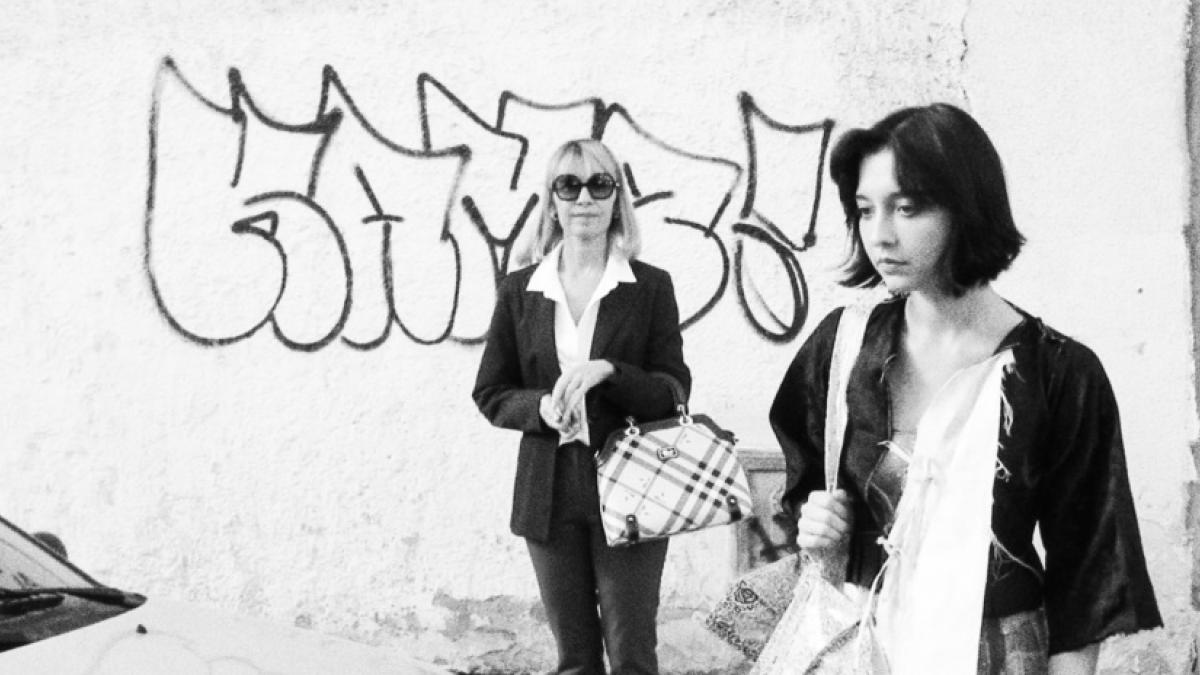 Black and white medium shot of two stylishly-dressed women starting to cross a street in front of a wall with graffiti.