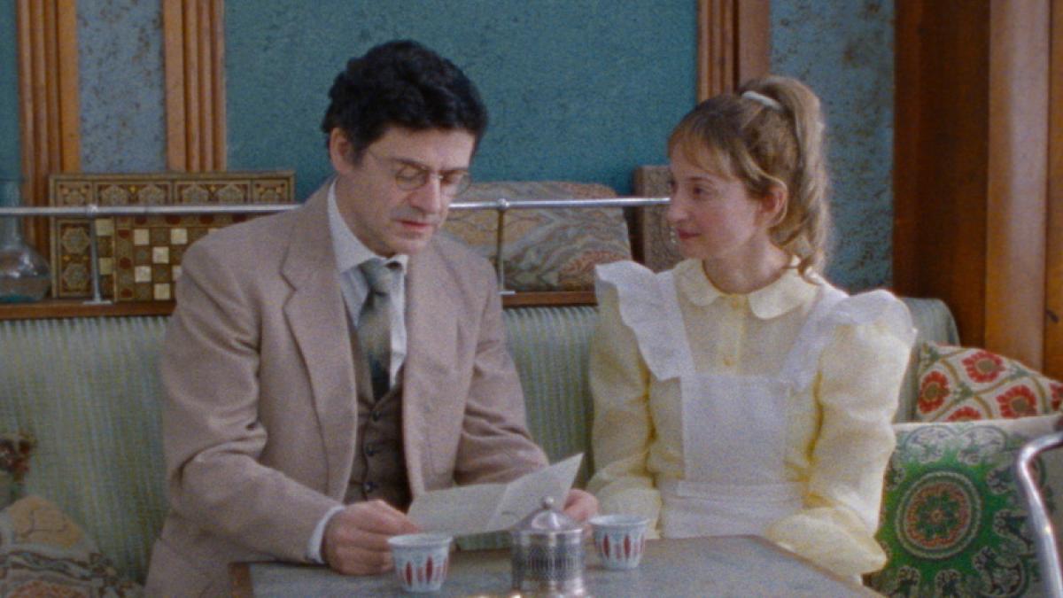 Medium shot of a man in a suit and a woman in a frilly dress sitting in a cafe.
