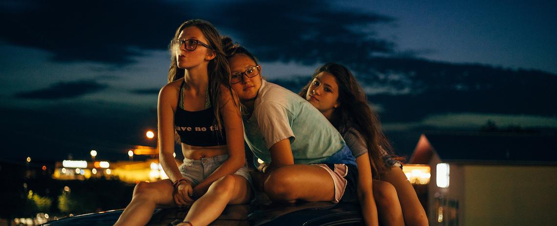 Three teenage girls sit together on the roof of a car shortly after sunset.