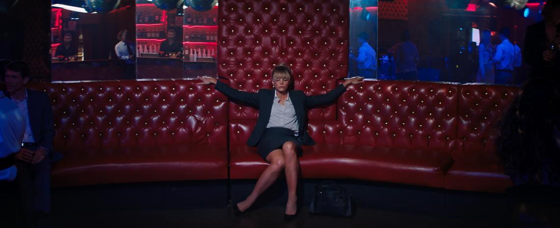 A young blond woman dressed in a business outfit with blouse, jacket, and skirt sits on a lounge seat in a bar, apparently half asleep.