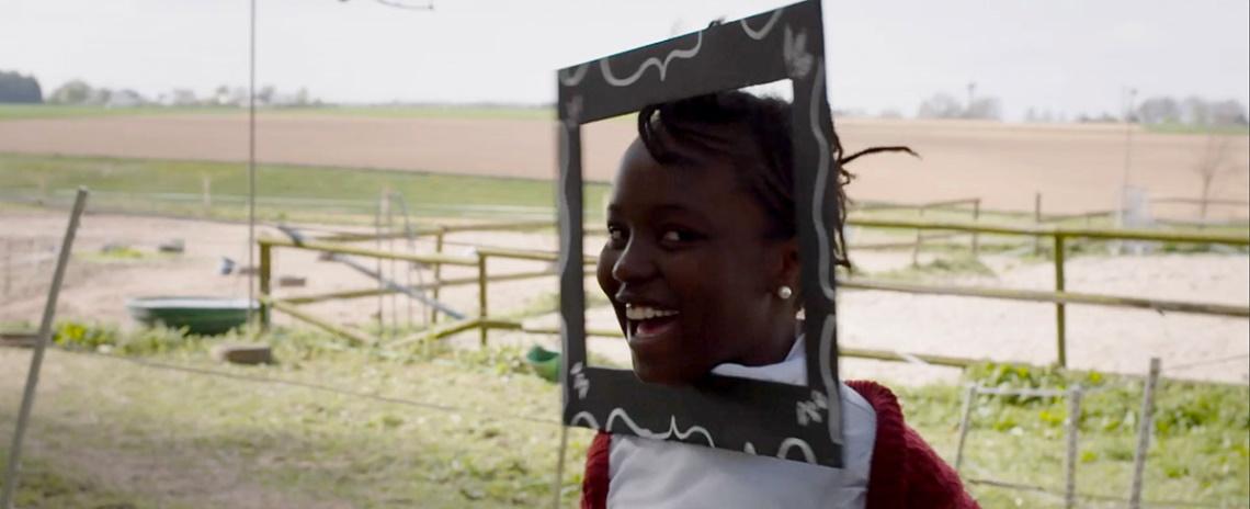 A laughing girl with a picture frame attached to her face, with fenced agricultural fields in the background.