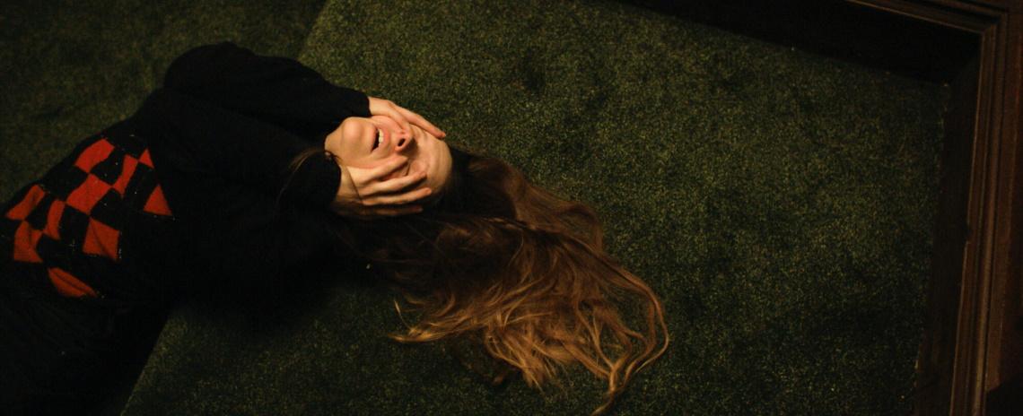 A woman in a black and red sweater lays on a green carpeted floor, her hands partly covering her face and her hair spread out on the ground.