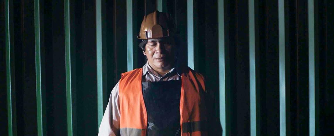 A middle-aged Native man in a hardhat, bulletproof vest, and safety vest stands in front of a shipping container, his eyes closed.
