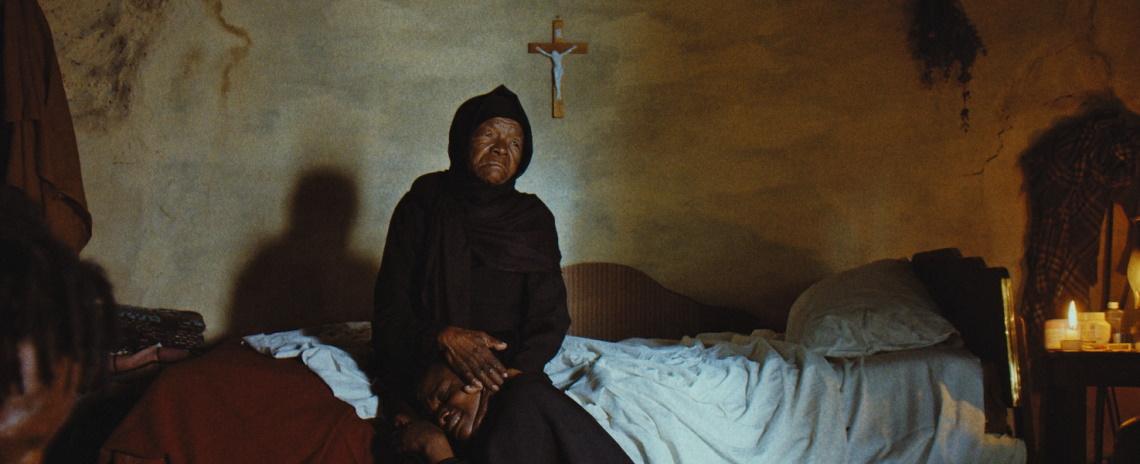 An elderly Black woman in long black robes and headscarf sits on a bed, cradling a younger Black man's head in her lap.