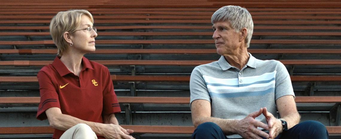 An older woman in a red polo shirt and older man in gray striped polo shirt sit side-by-side on sports bleachers, looking at one another.