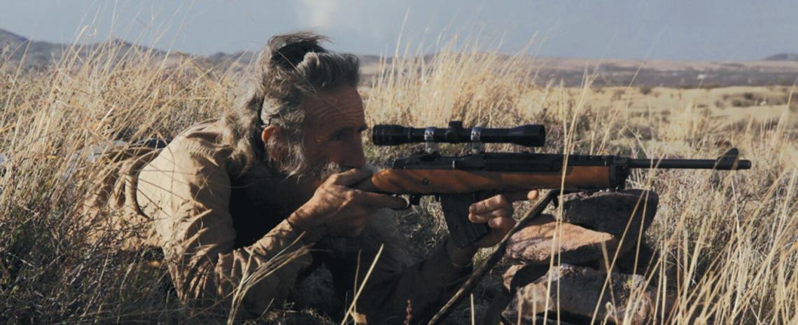 An older, bearded man laying down in a field of dry grass, pointing a rifle with a scope at something offscreen.