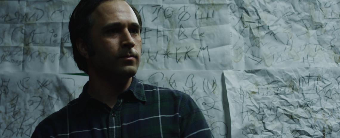 A dark-haired man standing in front of a wall covered in wrinkled sheets of paper, each with numerous scrawled symbols.