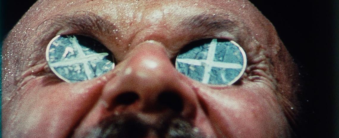 A close-up of a man's sweaty, upturned face. Two pennies painted with X's have been placed on his eyes.