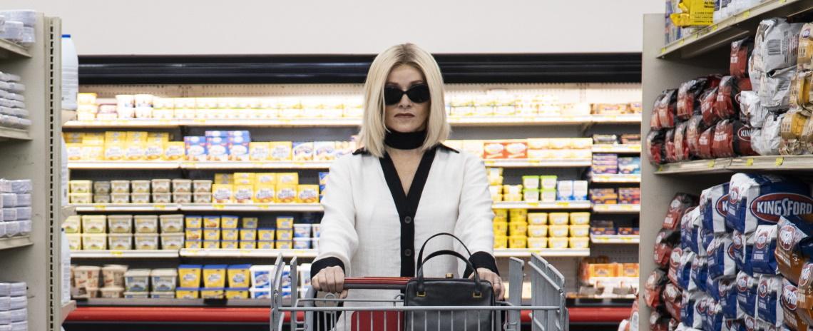 An elegant woman in a white coat in sunglasses pushing a shopping cart through a grocery store.