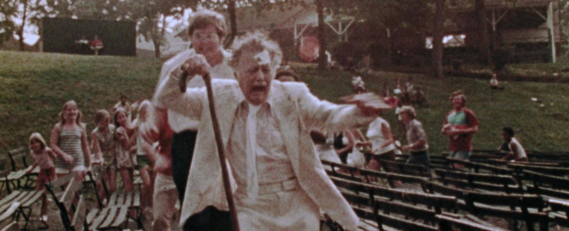 An elderly man in a white suit holding a cane flees in terror from a pursuing crowd of adults and children.