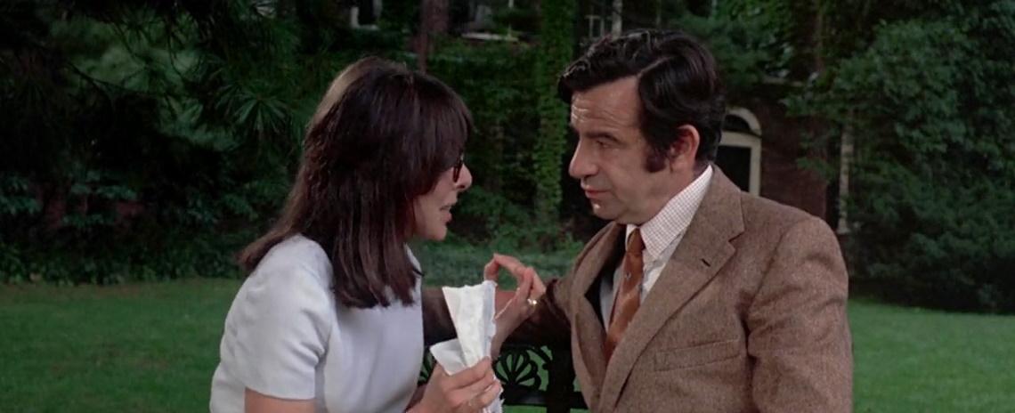 A woman in a white blouse and a man in a brown suit sit together, talking urgently, in a large, grassy outdoor area.