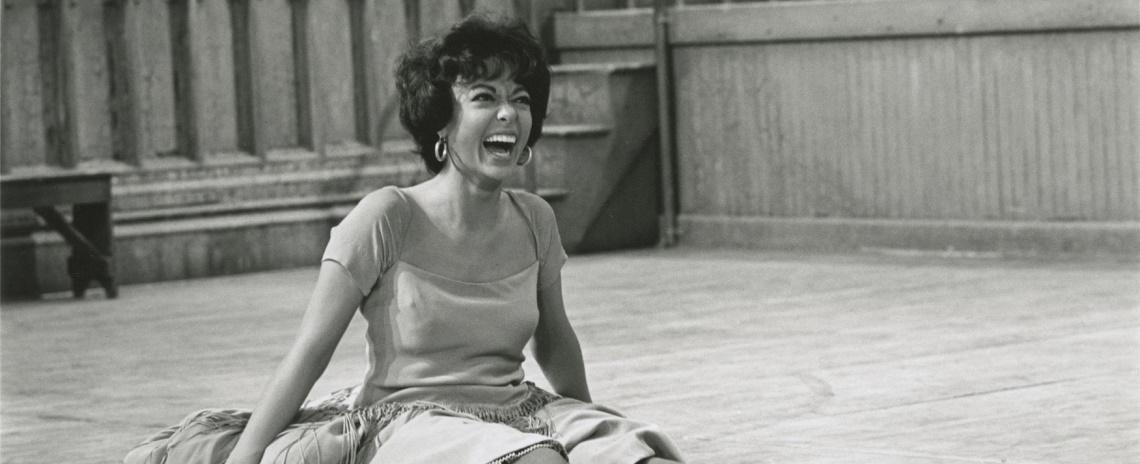 A black-and-white photo of a laughing, dark-haired woman in a dress, sitting on the floor of a dance studio.