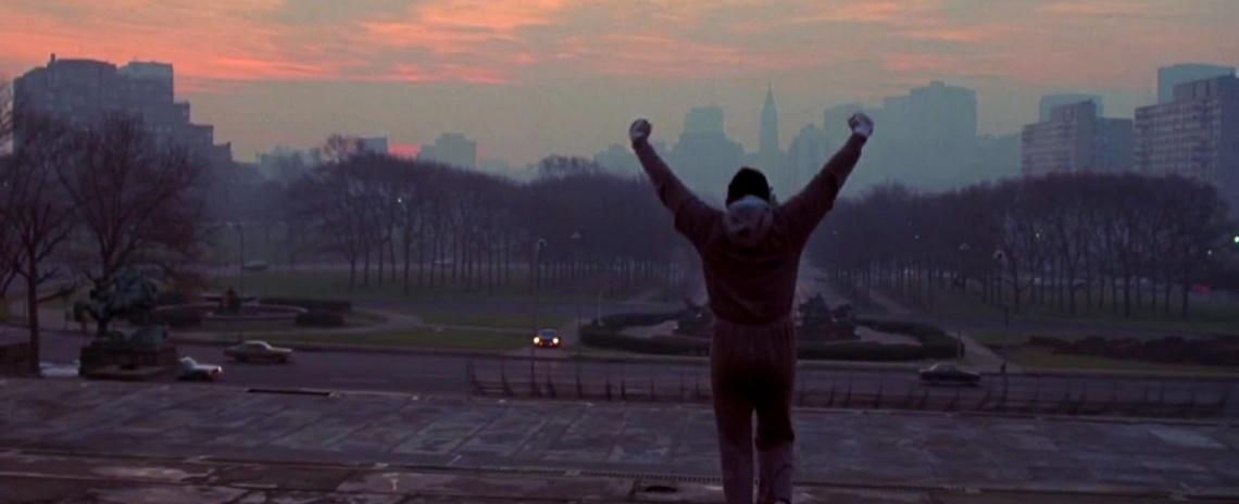 Wide shot of a man in sweatpants and hoodie, with his back to the viewer, holding his arms high in the air. In the background is a Philedelphia citscape at sunrise.