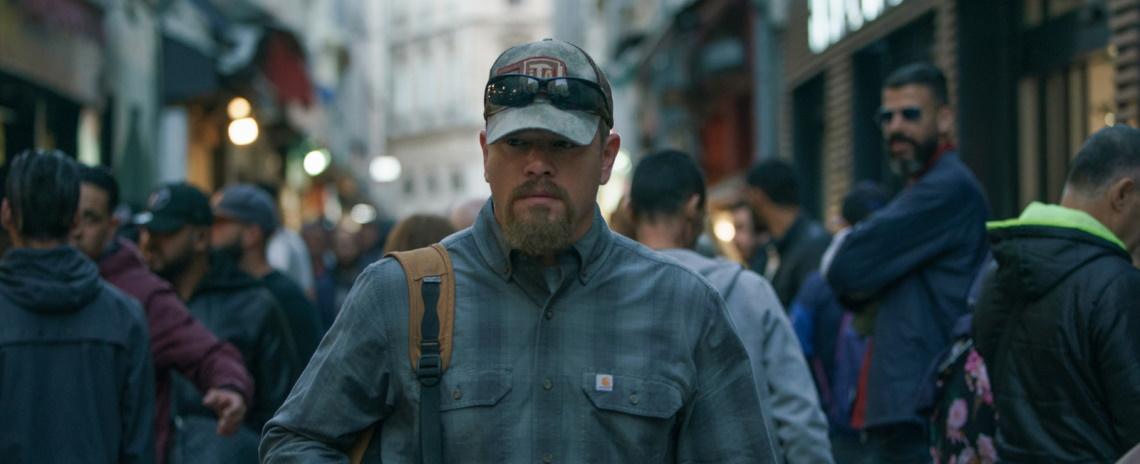 A man in a plaid workshirt and ball cap walks through a bustling French city.