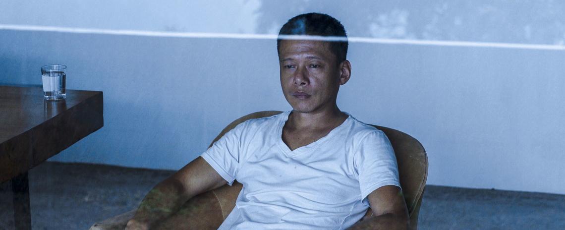 Medium shot of a man in a white t-shirt sitting in a chair in a spartan room, staring out the window at the falling rain.