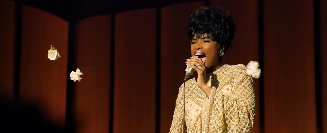 A medium shot of a Black woman in a vintage yellow gown and retro hairstyle singing into a microphone.