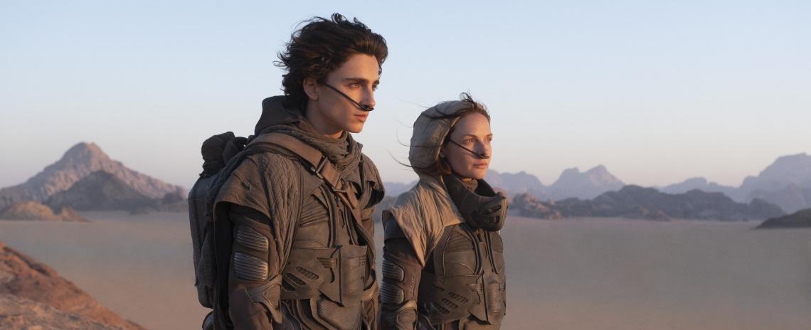Medium shot of a young man and a slightly older woman in futuristic jumpsuits, looking off to the right. Background is a barren desert landscape.